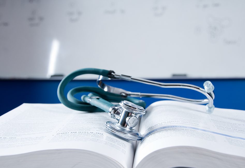 A steth rests on an open medical book in a classroom with a whiteboard behind it.