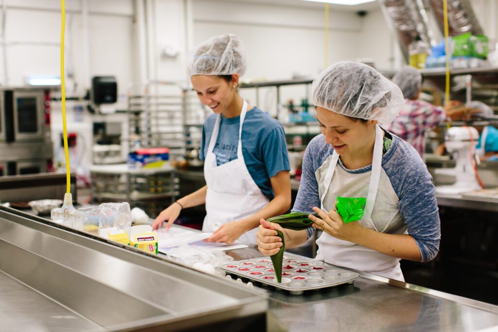 Two bakers stand side-by-side in a kitchen as one pipes cream into a cupcake tray.