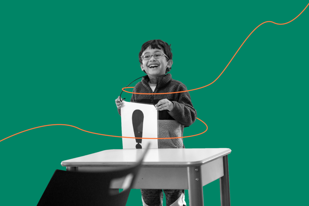 A smiling grade school student stands behind a desk in a public school and holds up a piece of paper with a large exclamation point printed on it.