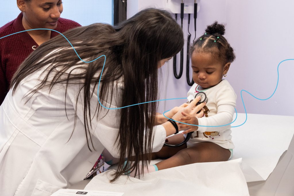 A doctor listens to a baby’s heartbeat through a stethoscope while a caregiver stands by.