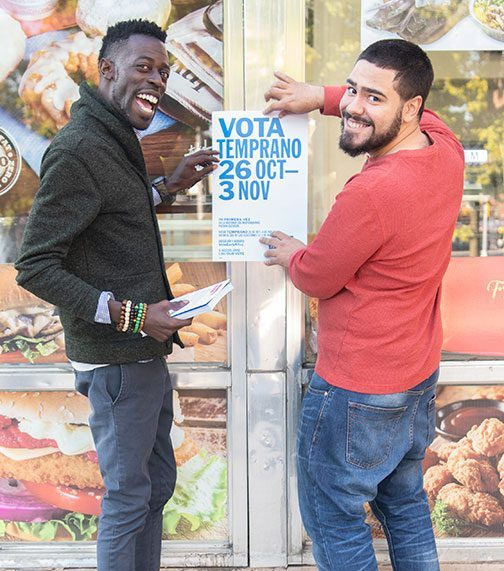 Two adults smile at the camera as they post a flyer to encourage voting.