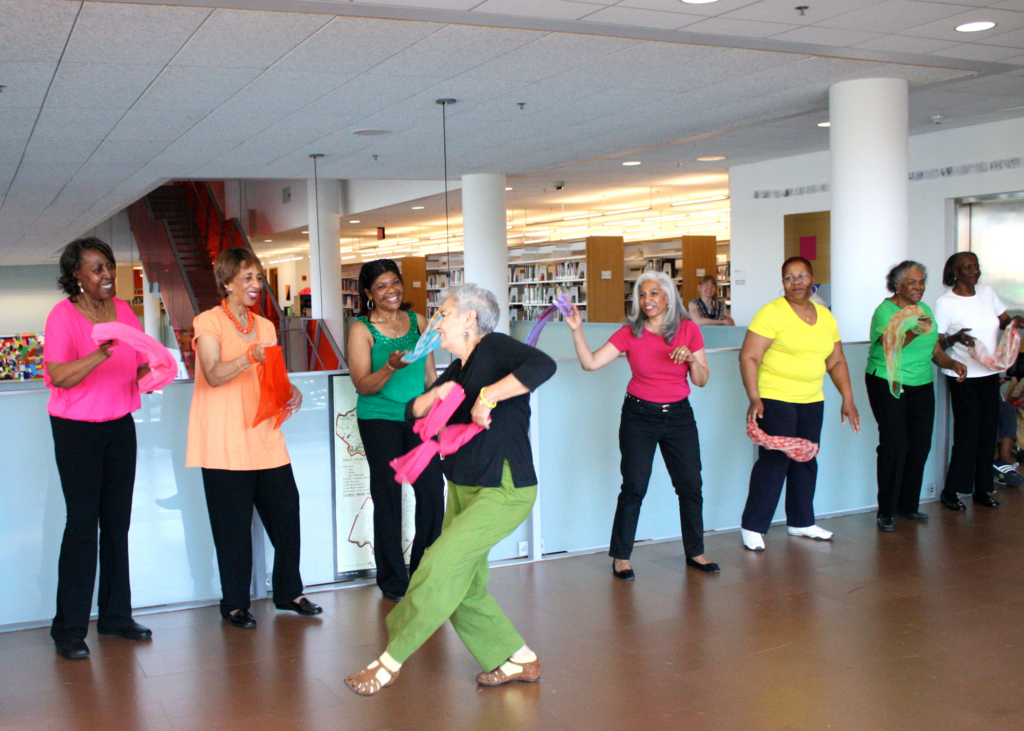 An older adult holding a bright pink scarf dances in front of five older adults who watch and smile while wearing colorful shirts and waving brightly colored scarves.