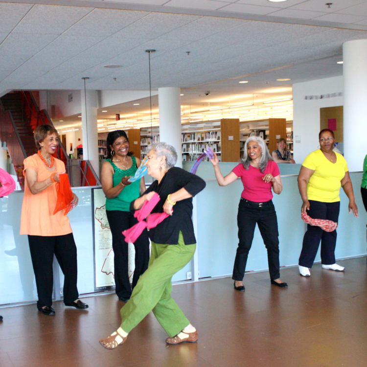 An older adult holding a bright pink scarf dances in front of five older adults who watch and smile while wearing colorful shirts and waving brightly colored scarves.