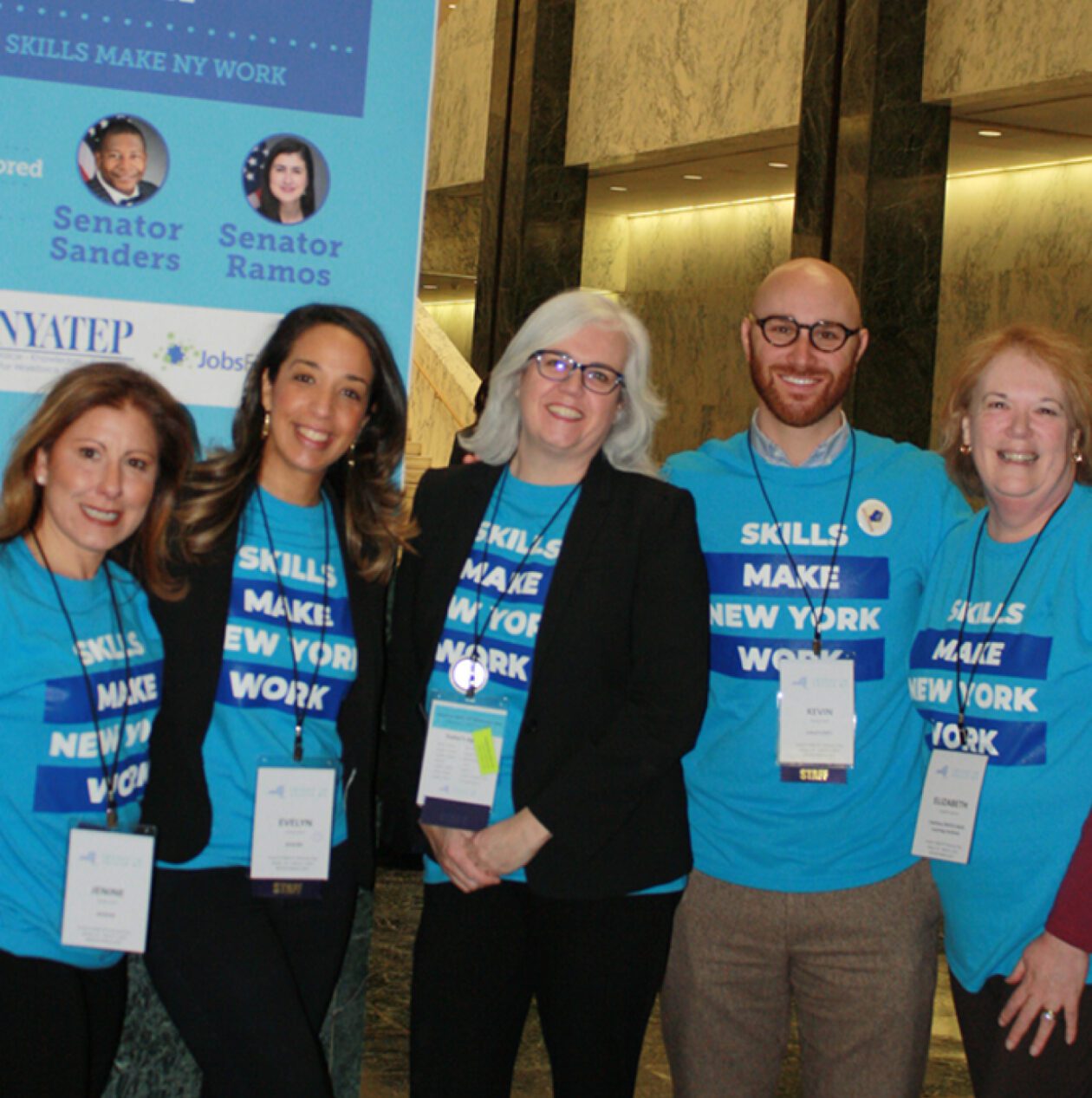 A group of advocates wearing tee-shirts that read “skills make New York work,” stand in a hall in Albany in front of a sign.