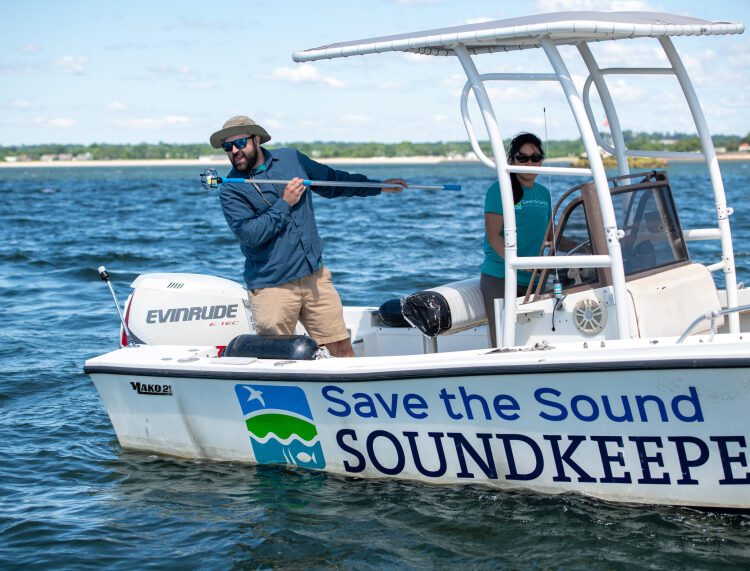 Two people on a boat collect water to assess its quality.