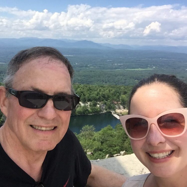 A man and a woman wearing sunglasses smile for the camera overlooking the Mohonk Preserve in upstate New York.