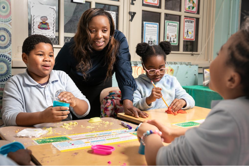 A woman and three children playing with Play Doh around school desks