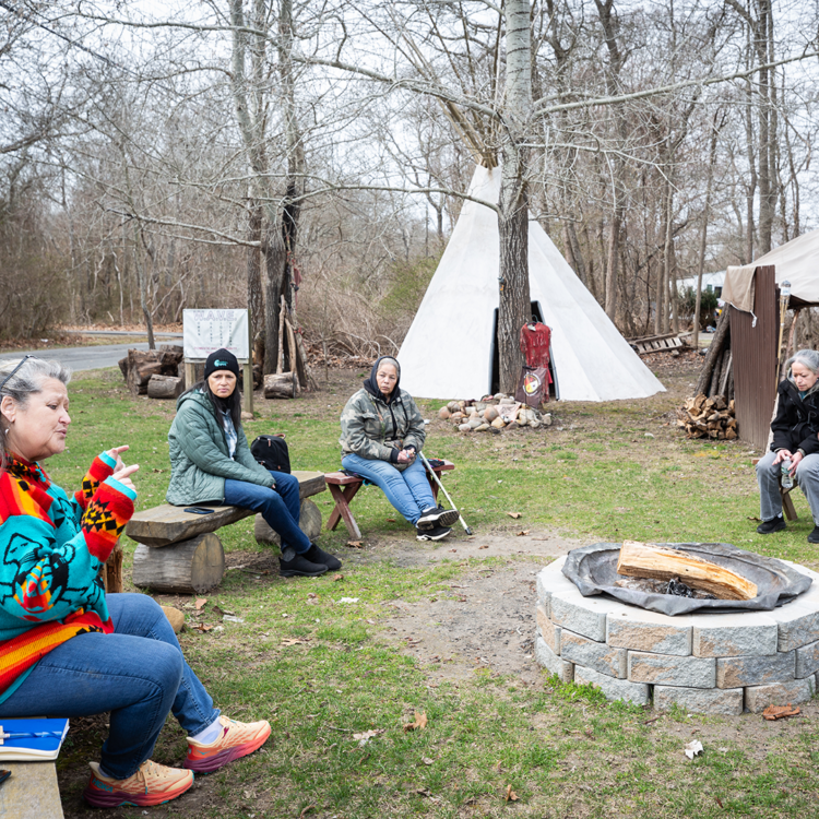 A group of people sit on wooden and stone benches around an unlit fire pit